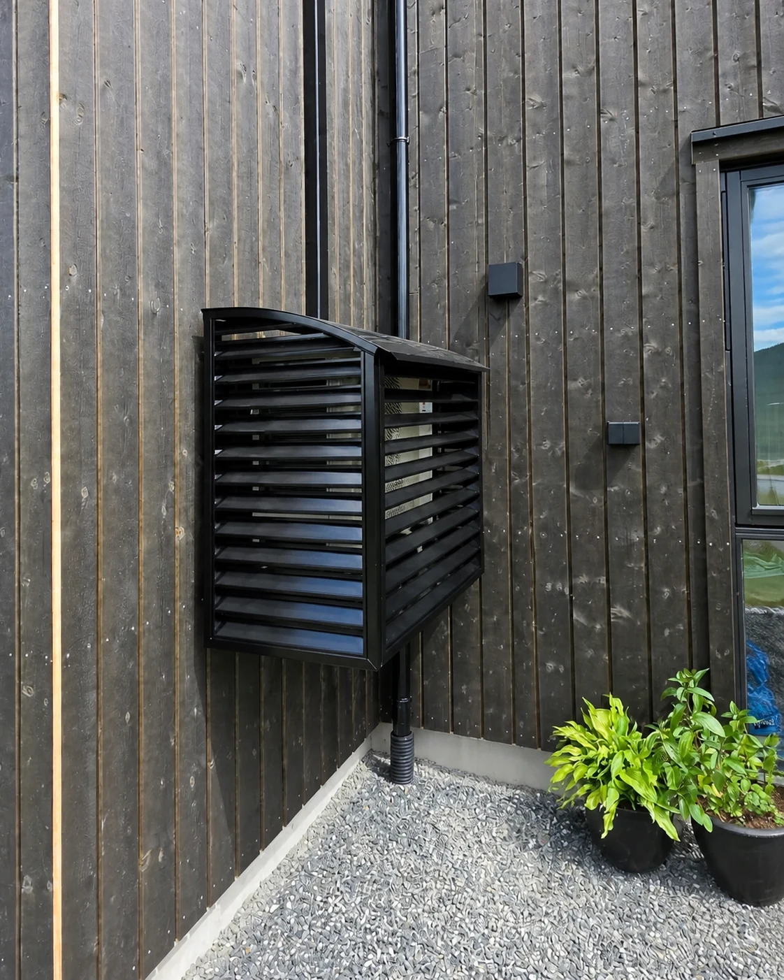 Sort varmepumpehus i lakkert metall med sort rørføring montert på mørk trevegg ved ny tomannsbolig på Lampeland i Flesberg